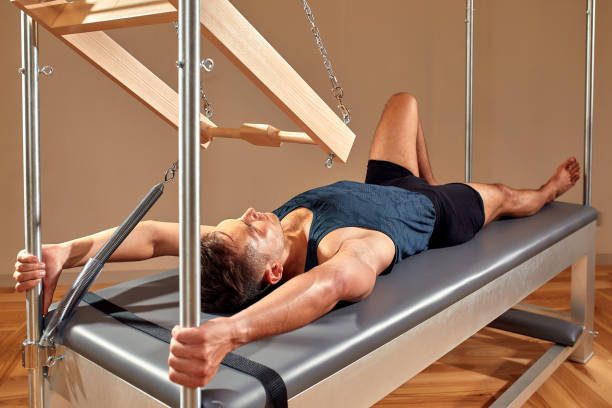 Side view of barefoot male athlete lying on pilates reformer and performing abs exercise during fitness workout. Pilates man concept